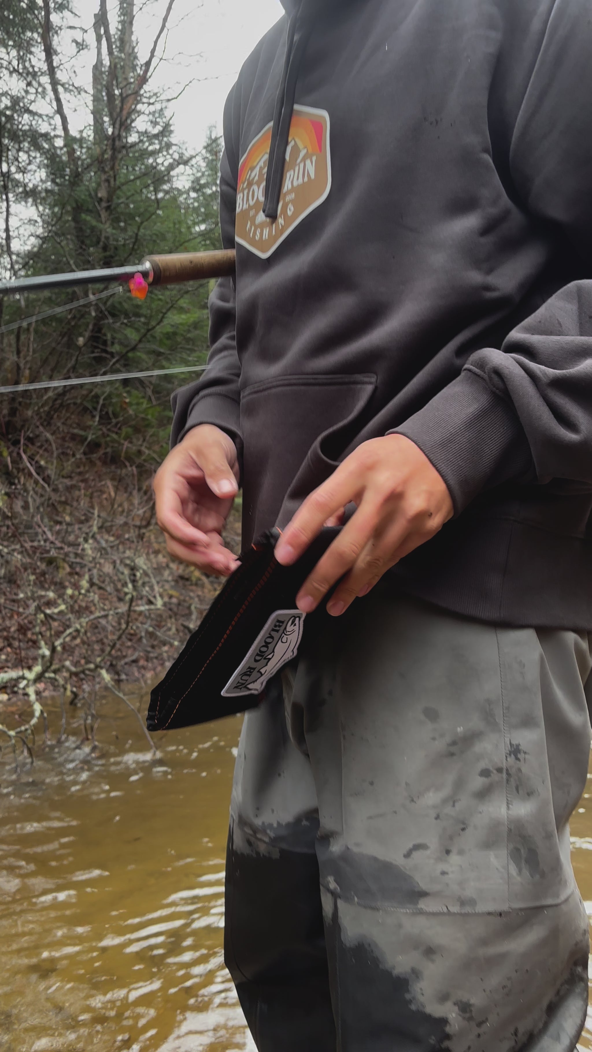 Caucasian male fishing for brook trout in a small stream in the Upper Peninsula of Michigan opening his Blood Run Fishing Black Float Fishing Wallet Bundle and removing a 5 gram camo Acorn fishing float wearing a men's coal black heavyweight fleece hoodie with the Blood Run Generator Logo on the chest and wearing a Blood Run men's brown camo knitted beanie.