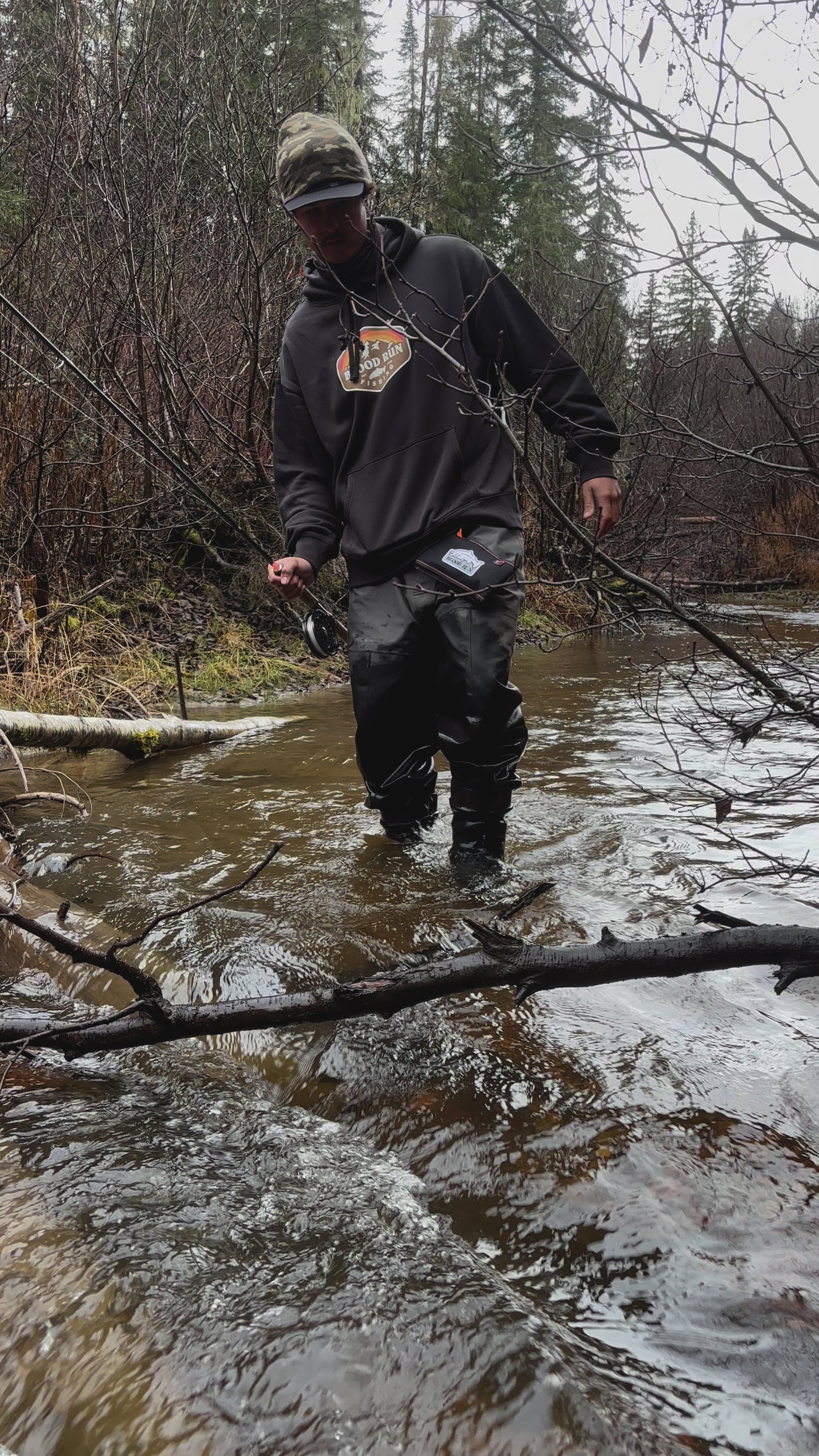 Caucasian male fishing for steelhead in a small stream in the Pacific Northwest using  a Blood Run Pinland 3 weight centerpin fishing rod, Halcyon 4.5" Centerpin reel, 15lb test clear Blood Run Float Fishing monofilament mainline, wearing a men's coal black heavyweight fleece hoodie with the Blood Run Sun Mountain Generator Logo on the chest and a brown camo knitted Blood Run Fishing beanie, carrying a Blood Run black float fishing wallet wearing simms waders.